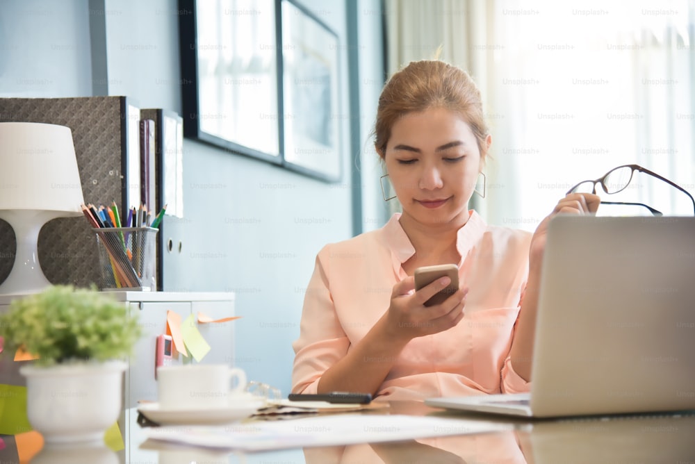 Junge asiatische Kreativdesignerin mit Handy an ihrem Arbeitsplatz. Eine Frau beim Online-Shopping mit Smartphone zu Hause. Laptop.