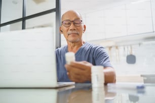 Homem adulto maduro asiático feliz segurando frasco de remédio enquanto consulta com o médico no laptop de casa, Telessaúde ou conceito de Telemedicina