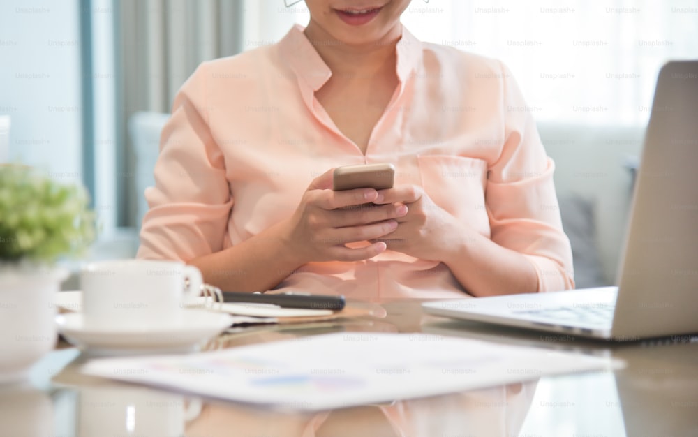 Joven diseñadora creativa asiática usando un teléfono celular en su lugar de trabajo. Una mujer haciendo compras en línea con un teléfono inteligente en casa. Ordenador portátil.