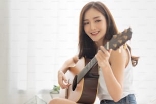 예쁜 아시아 여자가 복사 공간으로 기타를 연습하고 있다. 집의 하얀 방에서 악기를 들고 편안한 소녀.