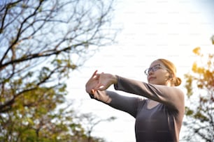 Happy Asian female in grey sportswear workout at the park. Young woman is warming up and stretching her arms at outdoors on morning. Health care and sport concept. Listening to the music with Earphones. Copy space. Exercising.