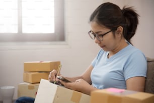 Close up Asian woman checking supplies and writing on cardboard box with mobile phone in her small warehouse. Own business