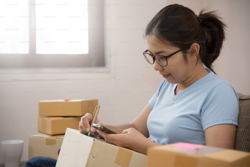 Gros plan sur une femme asiatique vérifiant les fournitures et écrivant sur une boîte en carton avec un téléphone portable dans son petit entrepôt. Entreprise propre