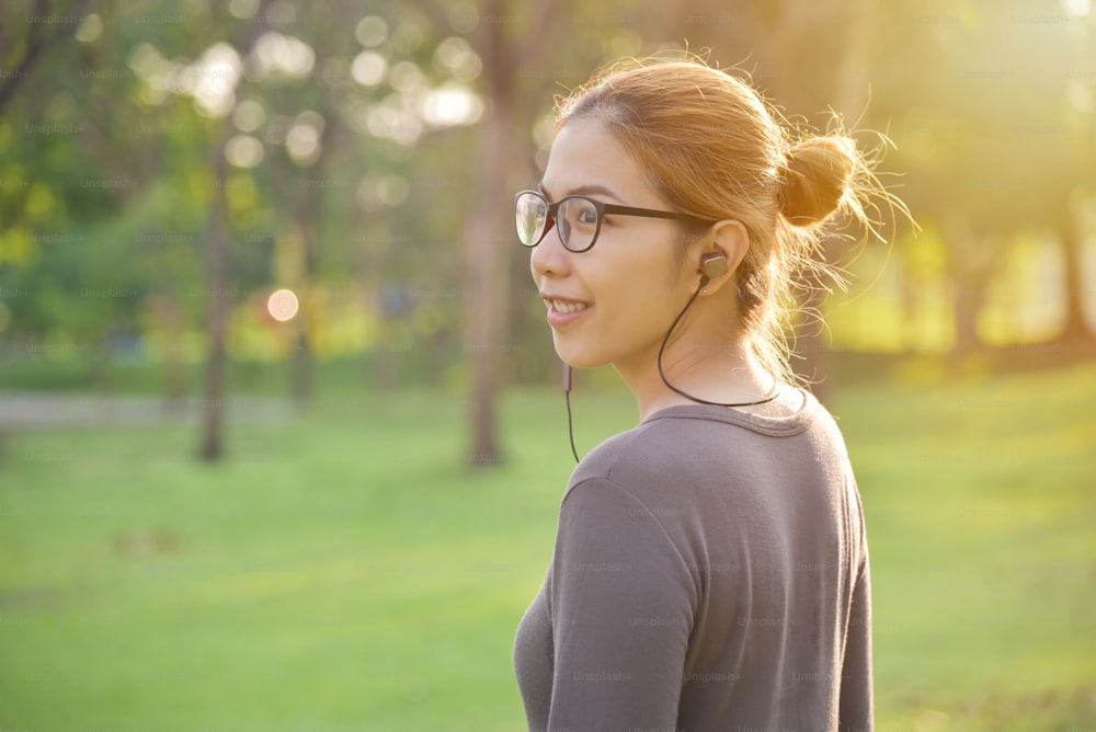 Une jeune femme asiatique heureuse en vêtements de sport gris sourit et écoute de la musique avec des écouteurs à l’extérieur le matin. Entraînement féminin au parc. Concept de soins de santé. Espace de copie.