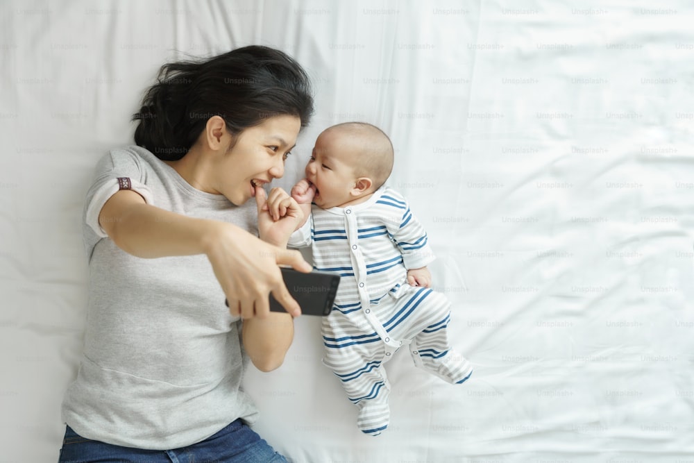 Asian Young mother and cute newborn baby playing together and using smartphone taking selfie on bed.