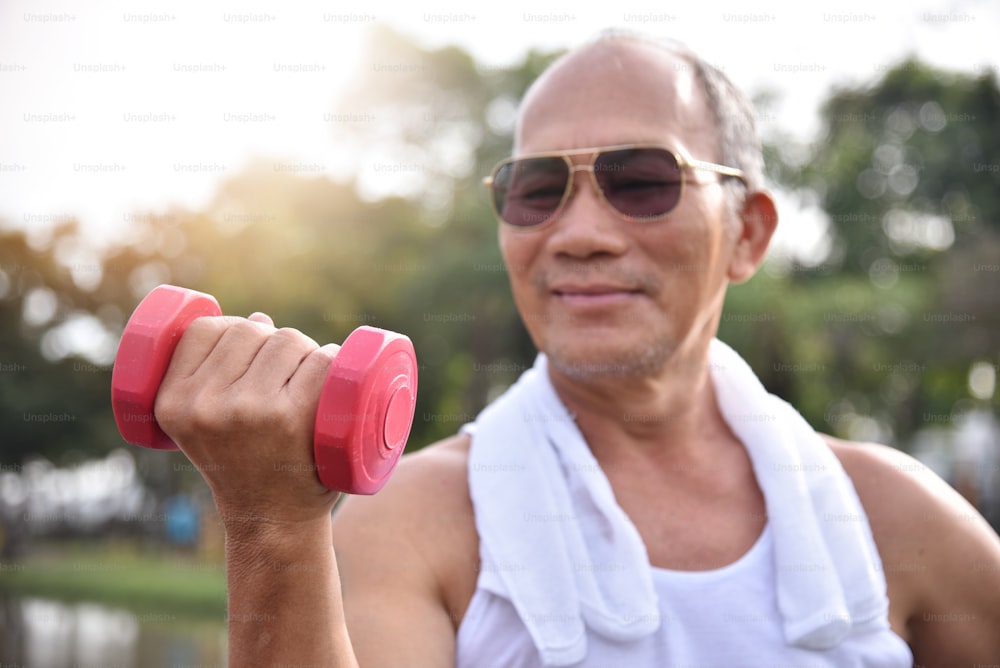 Glücklicher asiatischer älterer Mann trägt eine Sonnenbrille, die mit Hanteln im Park im Freien trainiert.