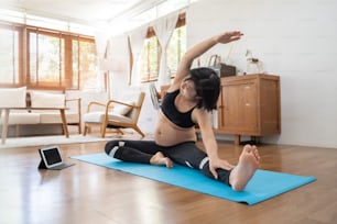 Asiatische schwangere Frau, die einen Online-Kurs auf einem digitalen Tablet ansieht, während sie zu Hause Yoga-Übungen macht. Schwanger weiblich, gesunde Schwangerschaft.