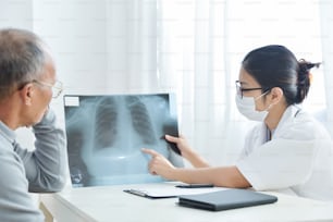 Young Asian female doctor with protective mask examining x-ray film of Senior male patient.