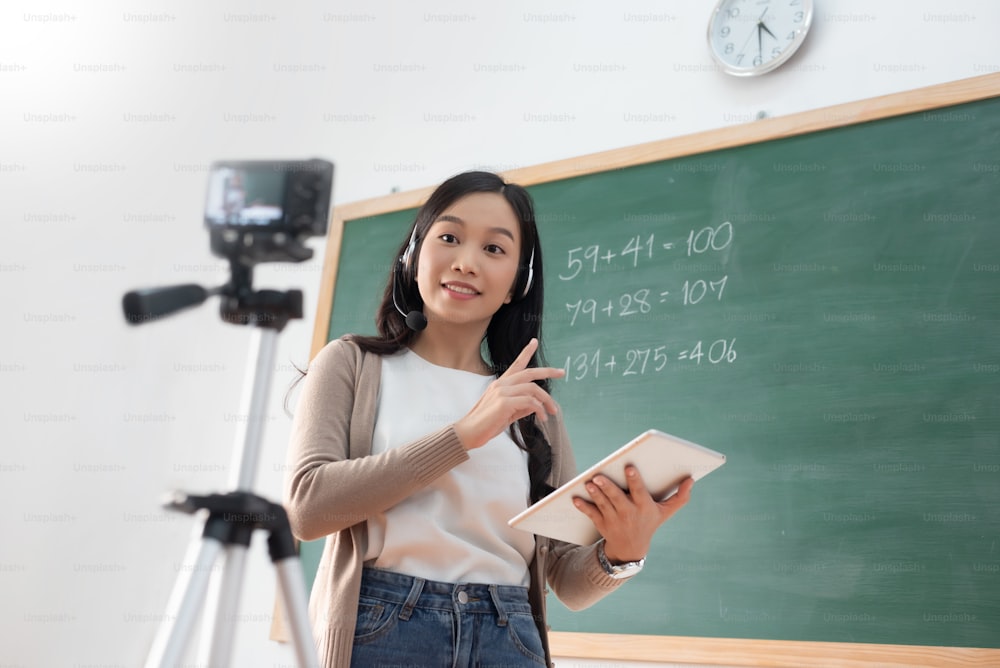 Chinese Woman teacher using digital camera, headphone and digital tablet standing online teaching in classroom at school