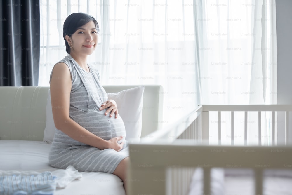 Ritratto di donna incinta asiatica, cinese o coreana sorridente felice seduta sul letto che tiene e tocca la pancia in camera da letto.