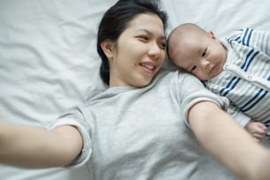 陽気なアジアの若い母と息子がベッドに横たわり、自宅で携帯電話で自撮りをしています。幸せな笑顔のお母さんと生まれたばかりの赤ちゃんが一緒にスマートフォンで写真を撮るのを楽しんでいます。愛する家族