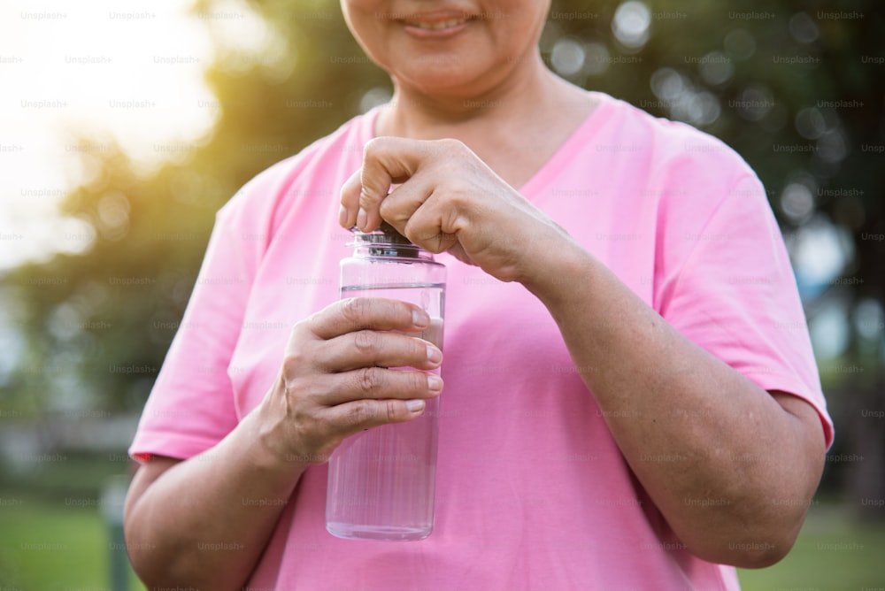 Mujer asiática mayor en camisa rosa sosteniendo una botella de agua para beber mientras hace ejercicio en el fondo al aire libre del parque.