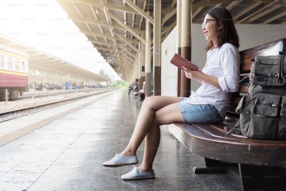 Mochileira asiática feminina está sentada na estação de trem tailandesa. A Jovem está lendo seu diário. Desfrutar de viagens.
