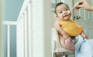 Asian little baby boy wearing yellow bib eating blend food on high chair at home.