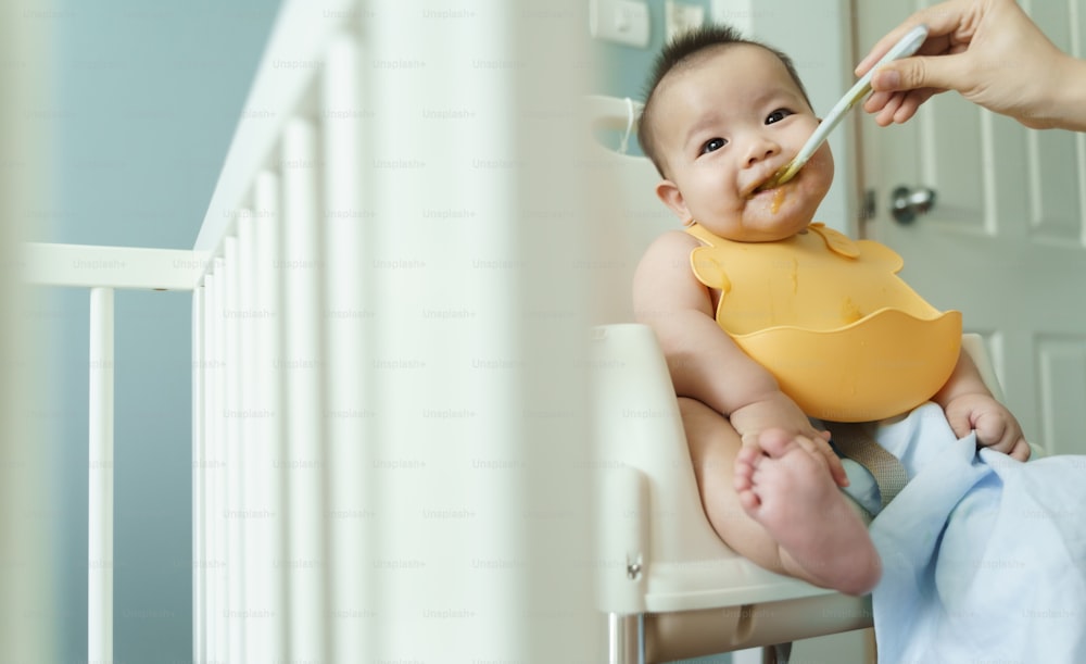Bebé asiático con babero amarillo comiendo comida mezclada en una silla alta en casa.