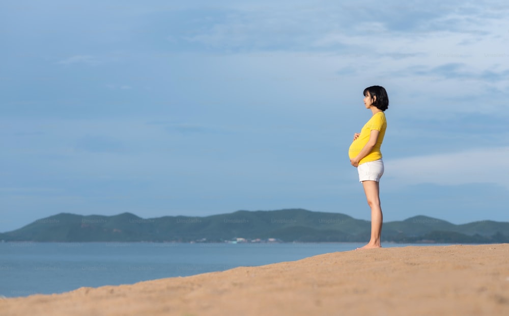 Relaxante asiático Mulher grávida em pé respirando ar fresco e segurando tocando sua barriga na praia ao ar livre.