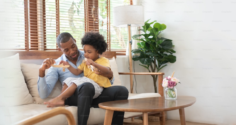 Padre afroamericano e hijo pequeño sentados en el sofá jugando juguetes de avión de madera en la sala de estar de casa juntos.