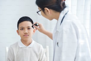 La joven doctora asiática usa anteojos que revisan los ojos de un paciente de niño pequeño con una linterna en el consultorio médico.