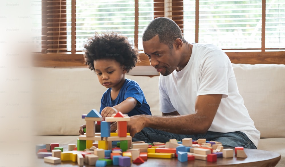 African Father and boy playing colourful wood blocks toy sitting on sofa at home.
