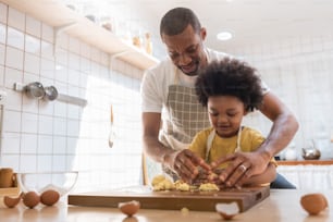 Padre afroamericano sonriente feliz enseñando al pequeño hijo amasando masa en la cocina, cocina de la familia negra o horneando galletas juntos en casa.