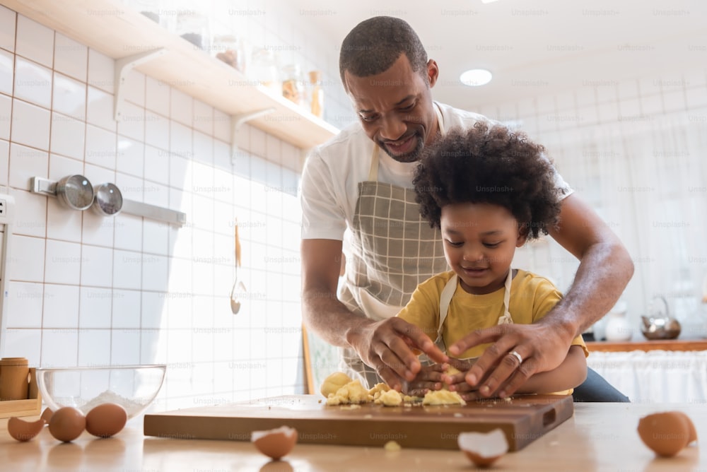 Happy Smiling African American Father teaching little Son kneading dough in kitchen, Black Family cooking or baking cookies together at home.