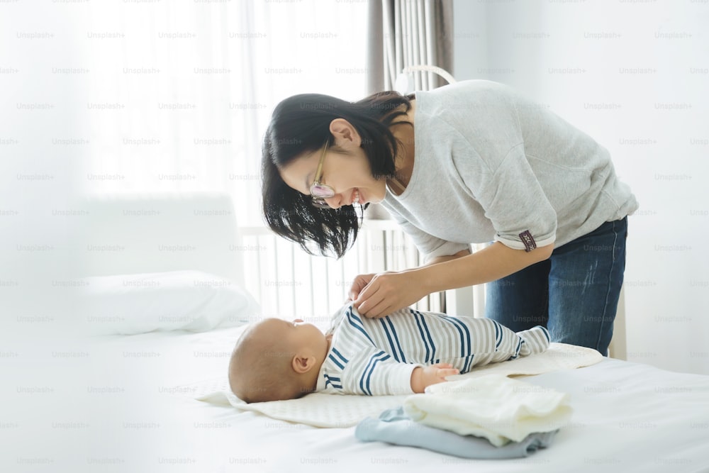 Loving Asian mother playing with little cute baby boy while dressing him on bed at home.