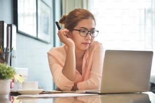 Young Asian Female creative designer wear glasses thinking and working with a laptop computer at her workplace. A Woman doing Online shopping at home.