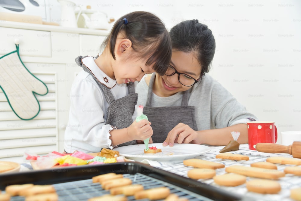 幸せなアジアの子供とキッチンでクッキーを飾る若い母親。