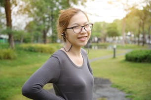 灰色のスポーツウェアを着た幸せな若いアジアの女性は、朝の屋外でイヤホンで笑顔で音楽を聴いています。公園での女性のトレーニング。ヘルスケアとスポーツのコンセプト。スペースをコピーします。行使。
