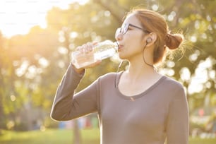 Young Asian female  in Grey sportswear is resting and drinking water while exercise at park. Woman listening to the music with earphones at outdoors on morning.  Health care concept.  Sunset and Sunlight. Copy space.