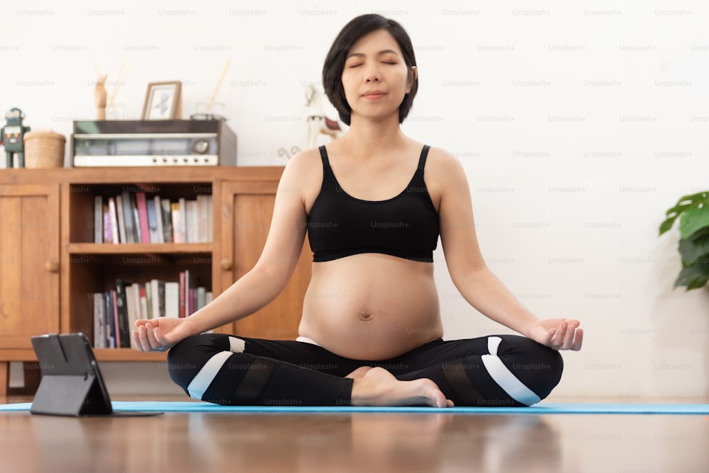 Jovem asiática mulher grávida meditando em yoga pose dentro de casa em casa, posição de lótus