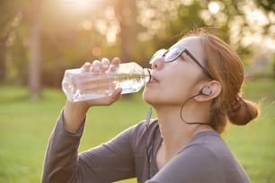 Una joven asiática en ropa deportiva gris está descansando y bebiendo agua mientras hace ejercicio en el parque. Mujer escuchando la música con auriculares al aire libre por la mañana.  Concepto de cuidado de la salud.  Atardecer y luz solar. Espacio de copia.