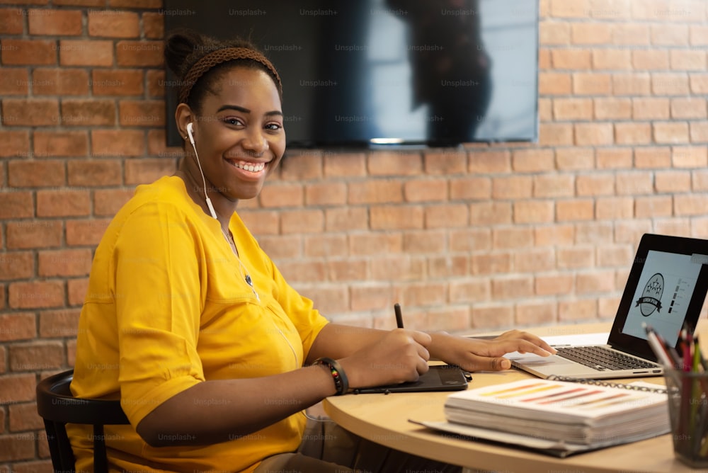 Portrait of African FeMale Creative Designer in yellow shirt with earphones sitting while using graphic tablet work on laptop computer.