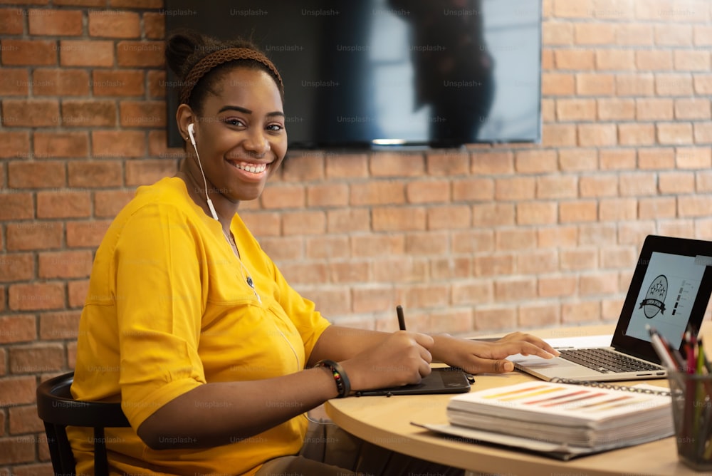 Retrato de un diseñador creativo afroafricano FeMale en camisa amarilla con auriculares sentado mientras usa el trabajo de la tableta gráfica en la computadora portátil.