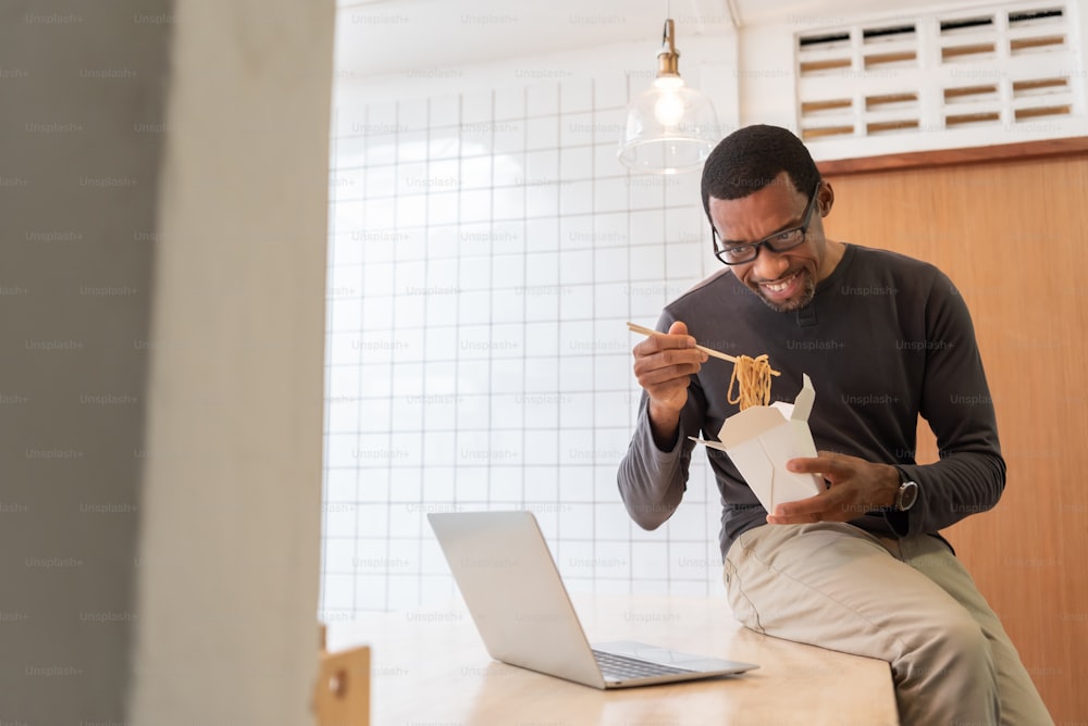 Uomo afroamericano che usa il computer portatile e che ha una chat video con i suoi amici mentre mangia una tazza istantanea di noodle a casa.
