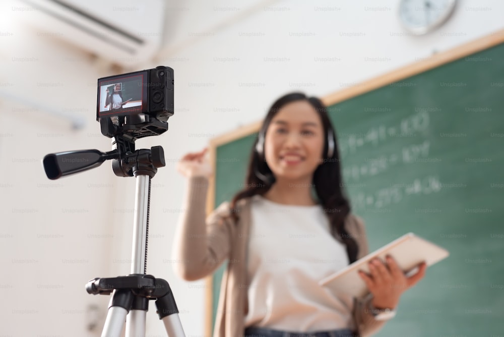 Enfoque selectivo, profesora joven asiática sonriente que enseña y graba video con cámara digital frente a la pizarra. Transmisión en vivo.