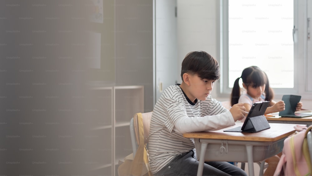 Concentrated Asian boy and girl sitting using digital tablet in classroom together at school. Education and Learning technology.