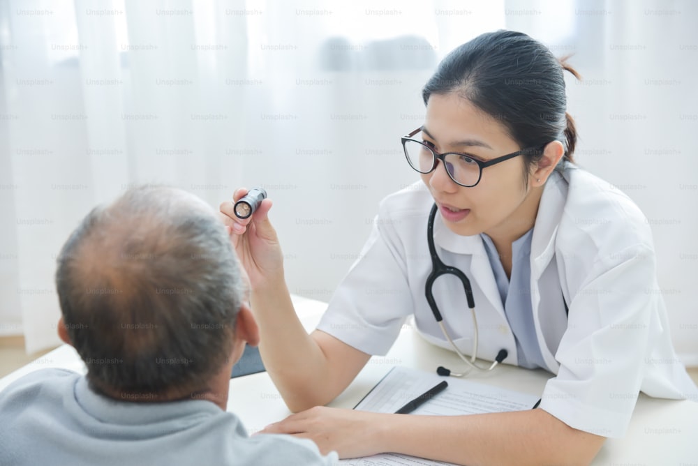 La joven doctora asiática usa anteojos que revisan los ojos del paciente del hombre mayor con una linterna en el consultorio médico.