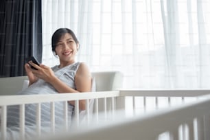Young beautiful Asian pregnant woman smiling using mobile phone in bed at home. Happy Expectant Chinese female holding smartphone and doing video chatting online with her family.