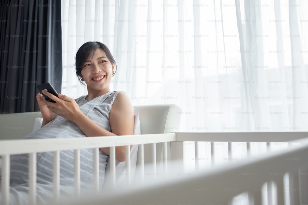 Giovane bella donna incinta asiatica sorridente usando il telefono cellulare a letto a casa. Felice femmina cinese in attesa che tiene lo smartphone e fa video chat online con la sua famiglia.