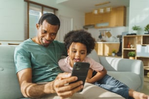 Padre e figlio afroamericani neri che usano lo smartphone a casa. Uomo africano sorridente felice e bambino piccolo che hanno videochiamata