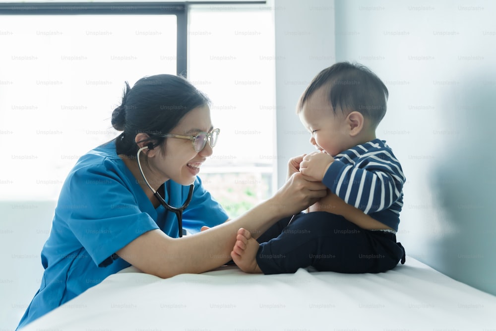 Fröhliche Chinesin oder Japanerin Ärztin spielt mit ihrer kleinen Patientin, asiatische Kinderärztin, die einen kleinen Jungen mit Stethoskop im medizinischen Untersuchungsraum untersucht
