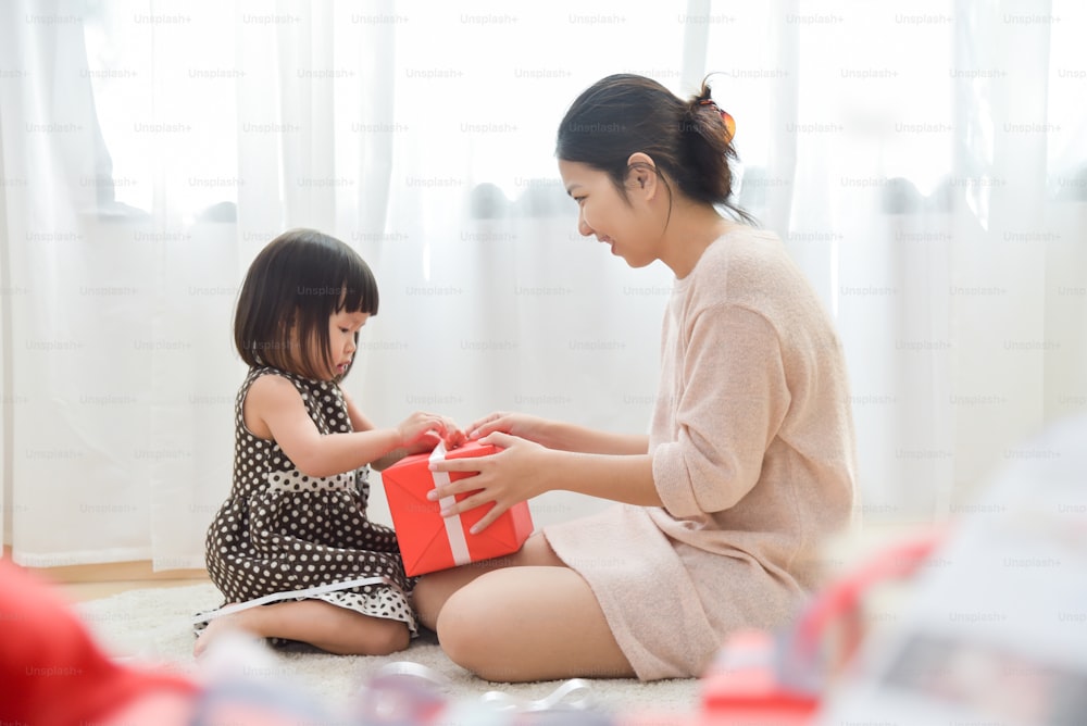 La joven madre asiática está mirando a una niña pequeña está desenvolviendo una caja de regalo roja en una habitación blanca para cumpleaños, Navidad y Año Nuevo. Espacio de copia. Feliz celebración en familia.