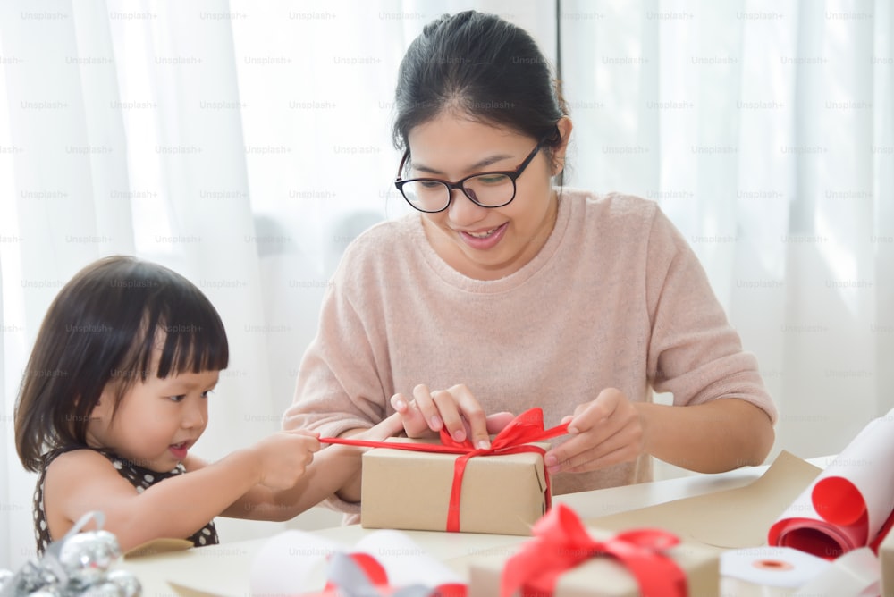 Joven madre y su hija envolviendo una caja de regalo para cumpleaños, Navidad y Año Nuevo en una habitación blanca. Familia asiática feliz en la casa. Espacio de copia.