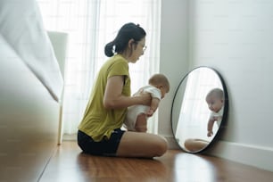 Madre asiática juguetona sosteniendo a su bebé recién nacido sentado mirando al espejo en casa. Alegre mamá joven sonriendo abrazando jugando con su hijo en el suelo juntos.