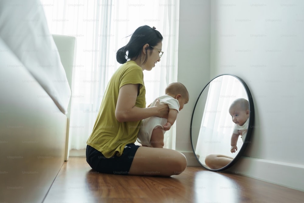 Playful Asian mother holding her newborn baby boy sitting looking at the mirror at home. Cheerful Young Mom smiling embracing playing with her son on the floor together.
