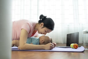 Feliz madre asiática deportiva joven haciendo ejercicio de plancha y jugando con su pequeño bebé en la colchoneta de yoga en casa. Mamá sana entrenando y practicando yoga con su adorable hijo en el suelo