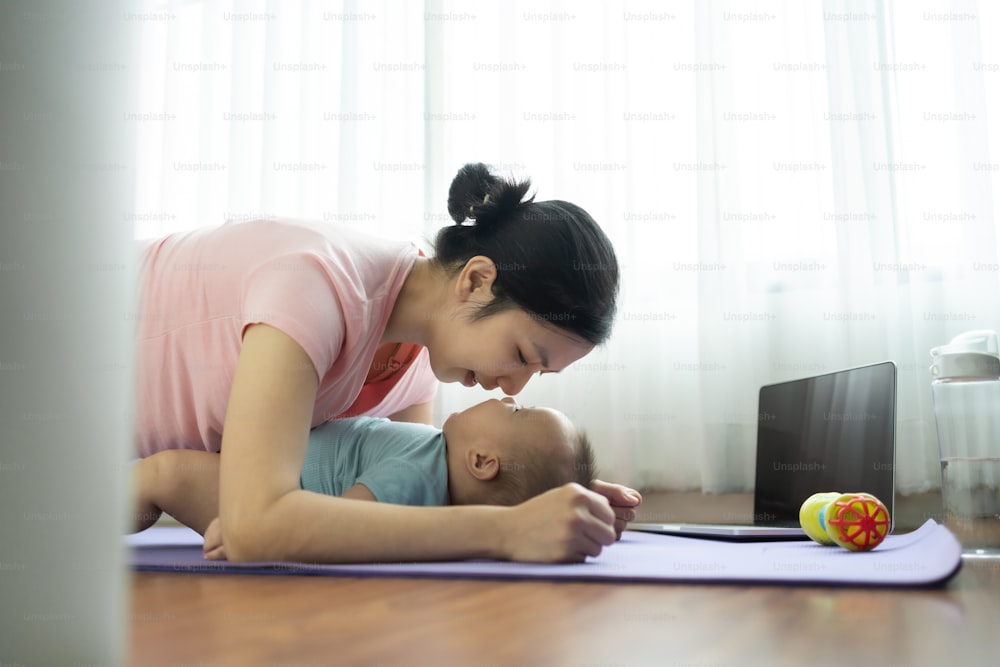 Happy Sporty Asian young mother doing plank exercise and playing with her little baby boy on yoga mat at home. Healthy Mom training and practicing yoga with her adorable son on the floor