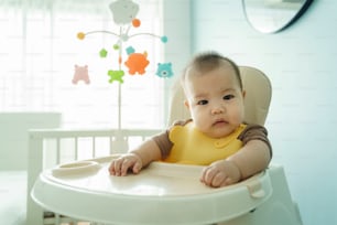 Retrato do menino pequeno bonito asiático sentado na cadeira alta enquanto espera por sua mãe alimentando a comida
