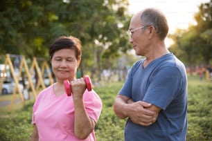 Asian Senior female exercising with lifting dumbbell while old man looking to her at park outdoor.